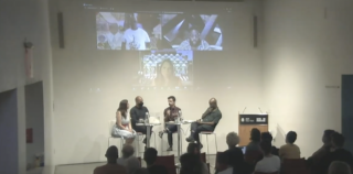 Four individuals sitting at the front of an auditorium with three individuals joining the conversation by zoom on the projected screen behind the four individuals