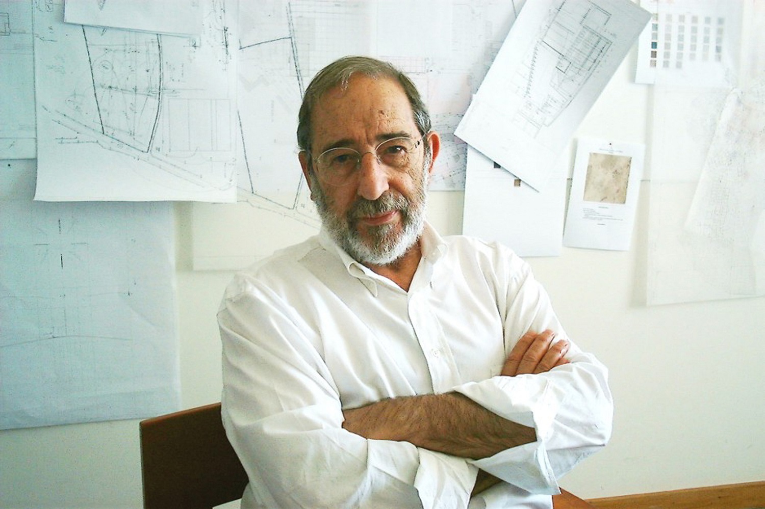 Álvaro Siza Vieira in a white shirt with building plans on a wall in the background