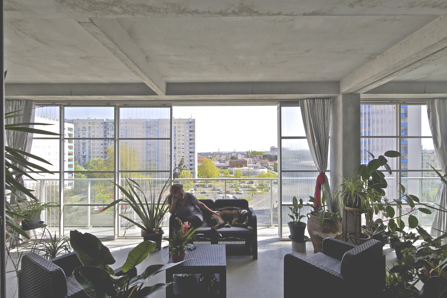 Photograph of an apartment in GRP Cite de Grand Parc showing a resident loungin on a couch with their dog in front of floor to ceiling windows