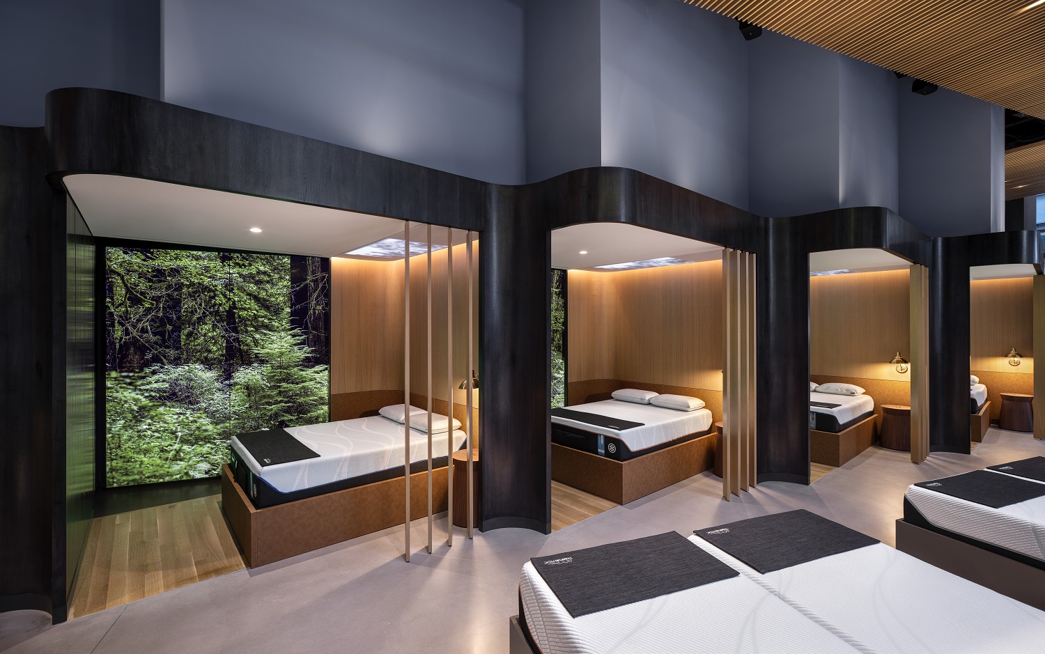 Photo of Tempur-Pedic showroom showing fours mattresses located in semi-private spaces on wooden floors with window-sized screens of a forest.