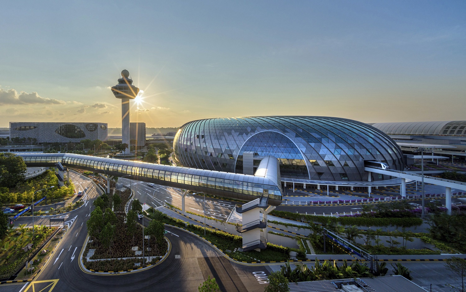 Exterior photo of Jewel Changi Airport, highlighting an all-glass bulbous building with a long pedestrian land bridge connecting to the structure