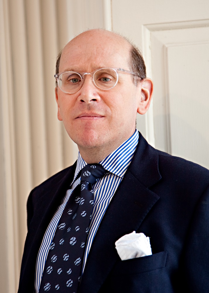 Headshot of Donalld Albrecht standing in a blue suit and tie and wearing clear glasses.