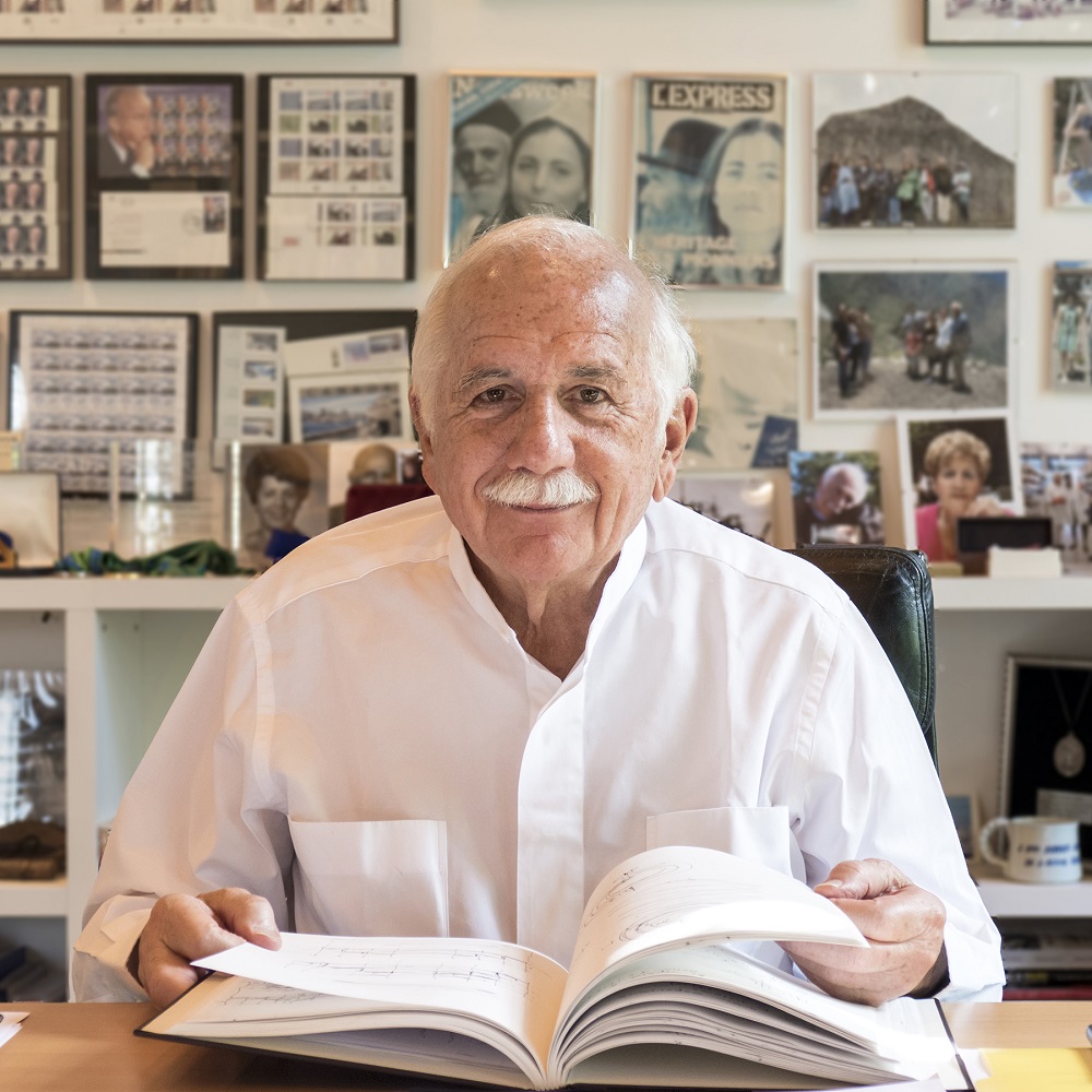 Headshot of Moshe Safdie wearing a white button down shirt sitting at a desk in an office while holding an open book. The background behind him is a wall covered in family and work photos.