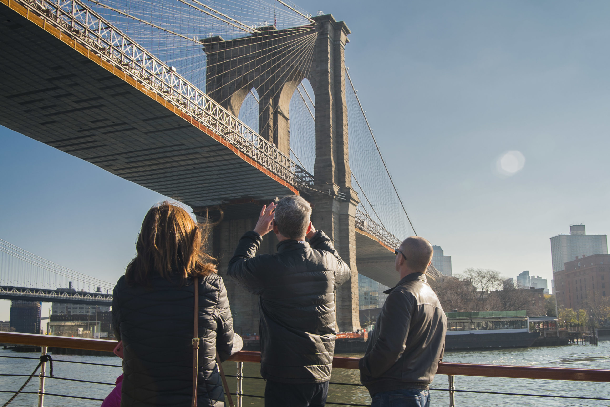The Brooklyn Bridge as seen from the deck of a Classic Harbor Line yacht with a few people standing in the foreground and taking photos of the bridge
