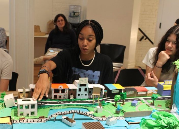 Middle school student gesturing towards a scale model of a neighborhood