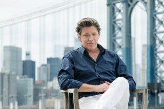 Headshot of Brendan Guerin, Co-founder and Principal, Guerin Glass Architects sitting in a chair with a backdrop of a bridge