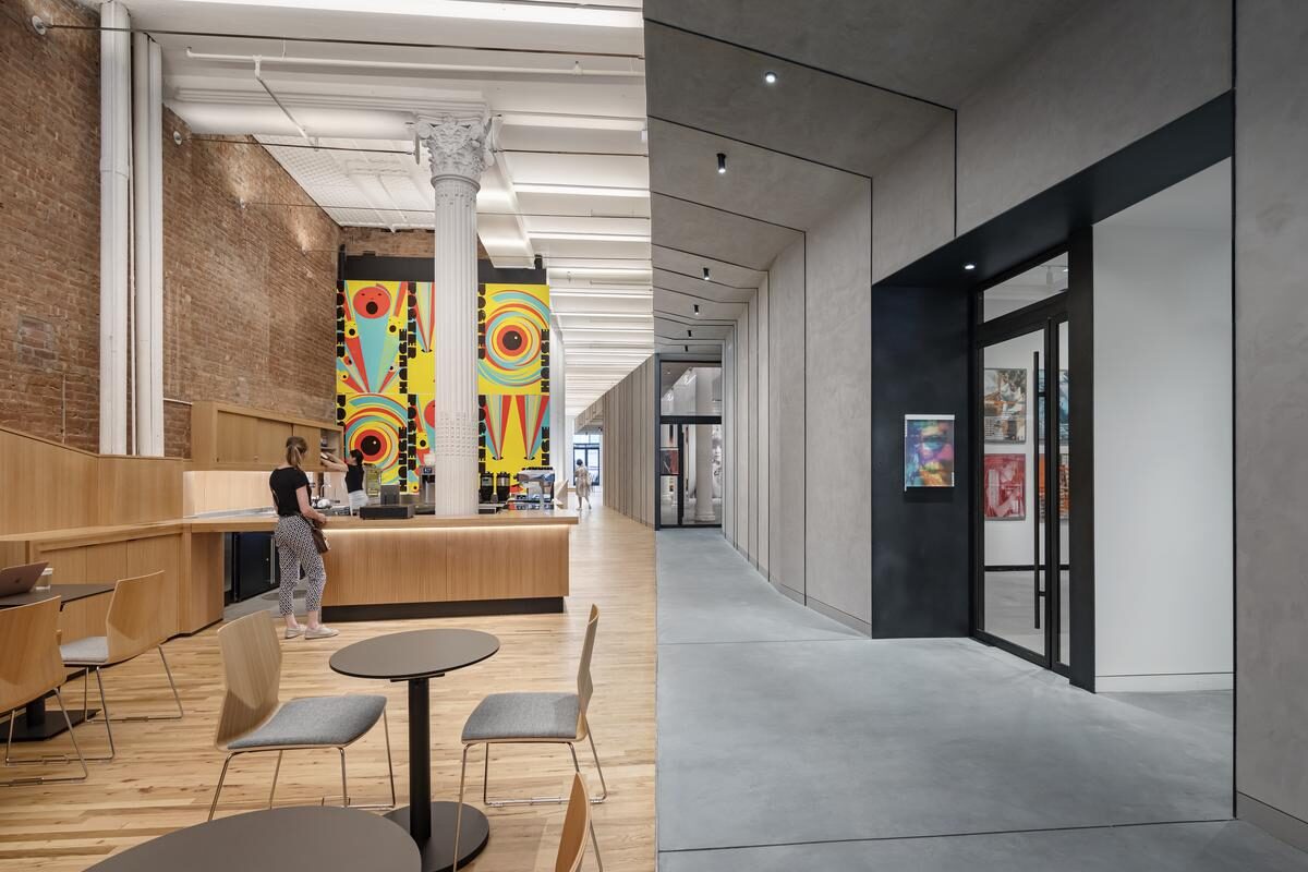 Interior of a building lobby with brightly lit warm colored materials, high ceilings and ornamental column on left and dimly lit dark grey monochromatic low ceilings on right.