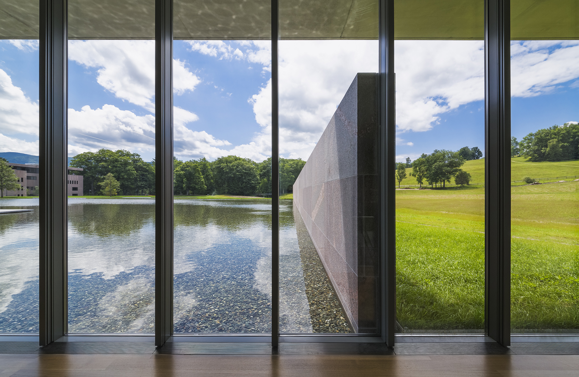 View looking out floor to ceiling windows with a reflection pool on the left and a green lawn on the right, separated by a single story wall