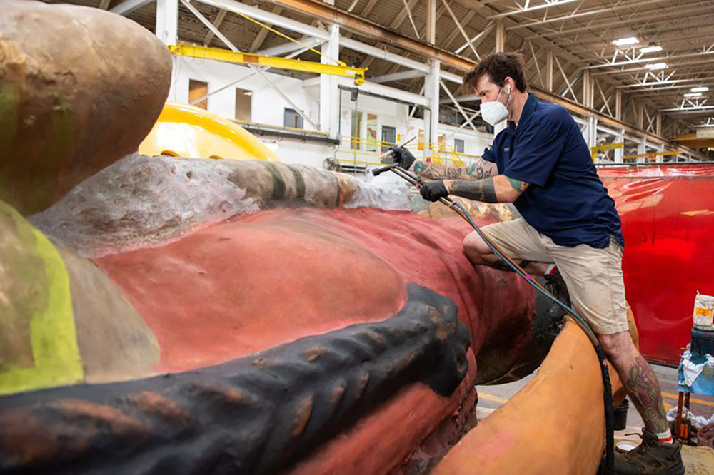 A man in a navy shirt and khaki shorts wearing a face mask holding a spray up to a large art sculpture taking up the foreground.