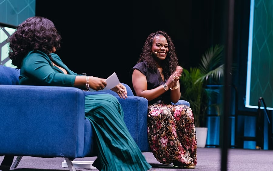 Two women seated on a stage on armchairs and holding microphones